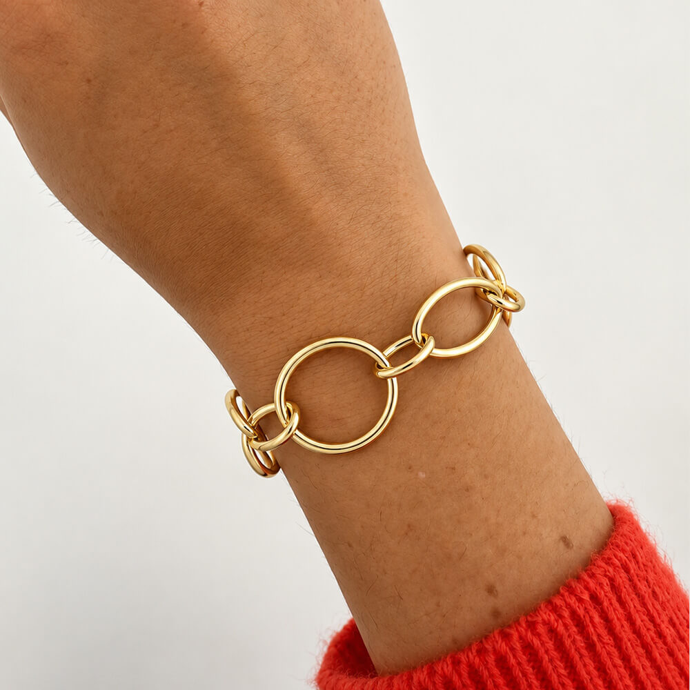 A close-up of an 18k yellow gold link bracelet with interlocking oval and circular links worn on a woman's wrist, with a red knit sweater cuff visible against a plain light background.