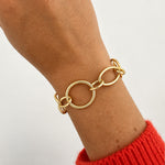 A close-up of an 18k yellow gold link bracelet with interlocking oval and circular links worn on a woman's wrist, with a red knit sweater cuff visible against a plain light background.