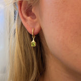 A close-up of a woman's ear wearing a small 18k gold huggie hoop earring with a dangling square-shaped green peridot gemstone.
