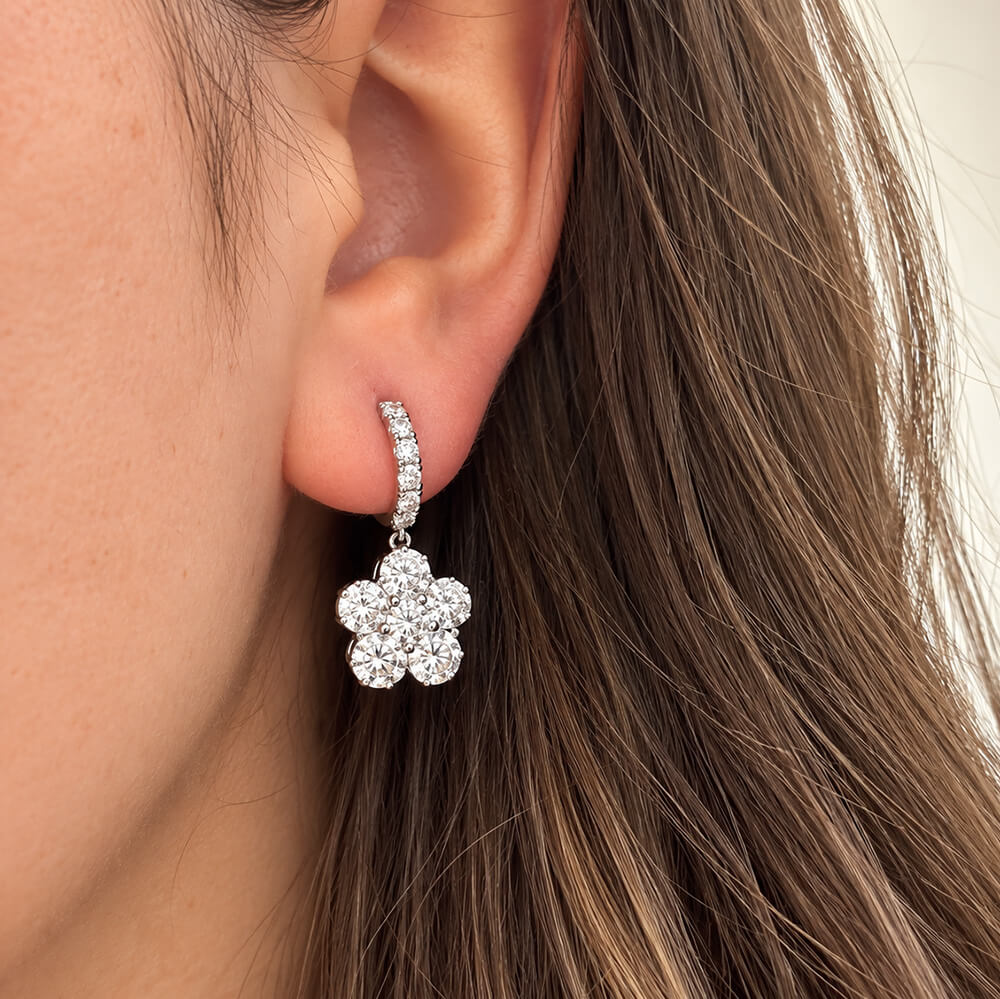 A close-up of a woman's ear wearing an 18k white gold huggie hoop earring adorned with small clear diamonds and a dangling flower-shaped cluster of clear diamonds.