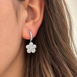 A close-up of a woman's ear wearing an 18k white gold huggie hoop earring adorned with small clear diamonds and a dangling flower-shaped cluster of clear diamonds.
