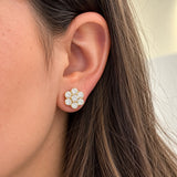 A close-up of a woman's ear wearing a flower-shaped stud earring made of clear diamonds in an 18k yellos gold setting.
