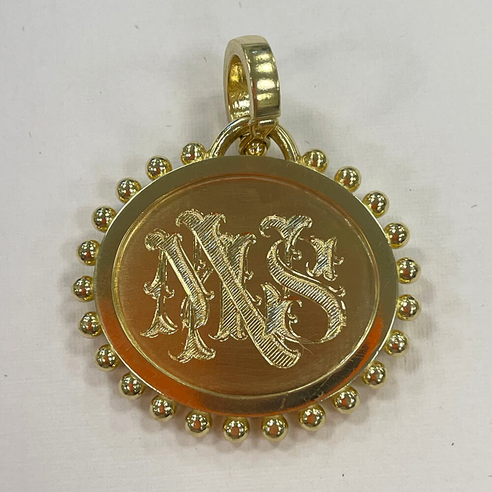 An 18K yellow gold round pendant featuring an engraved monogram 'MMS' in a gothic font, surrounded by a border of small gold beads, shown on a plain light background.