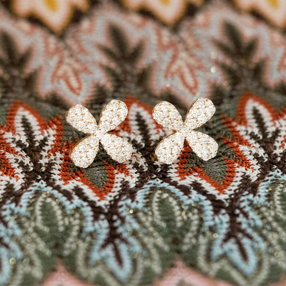 A pair of four-petal flower stud earrings covered in sparkling clear diamonds, resting on a colorful patterned knit fabric.