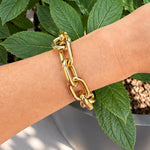 A close-up of a woman's wrist wearing a chunky 18K yellow gold chain link bracelet, set against a background of green leaves and a gray planter.