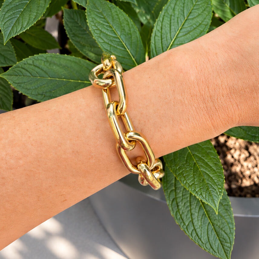 A close-up of a woman's wrist wearing a chunky 18K yellow gold chain link bracelet, set against a background of green leaves and a gray planter.