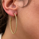 A close-up of a woman's ear wearing a thick, twisted 18k yellow gold hoop earring.