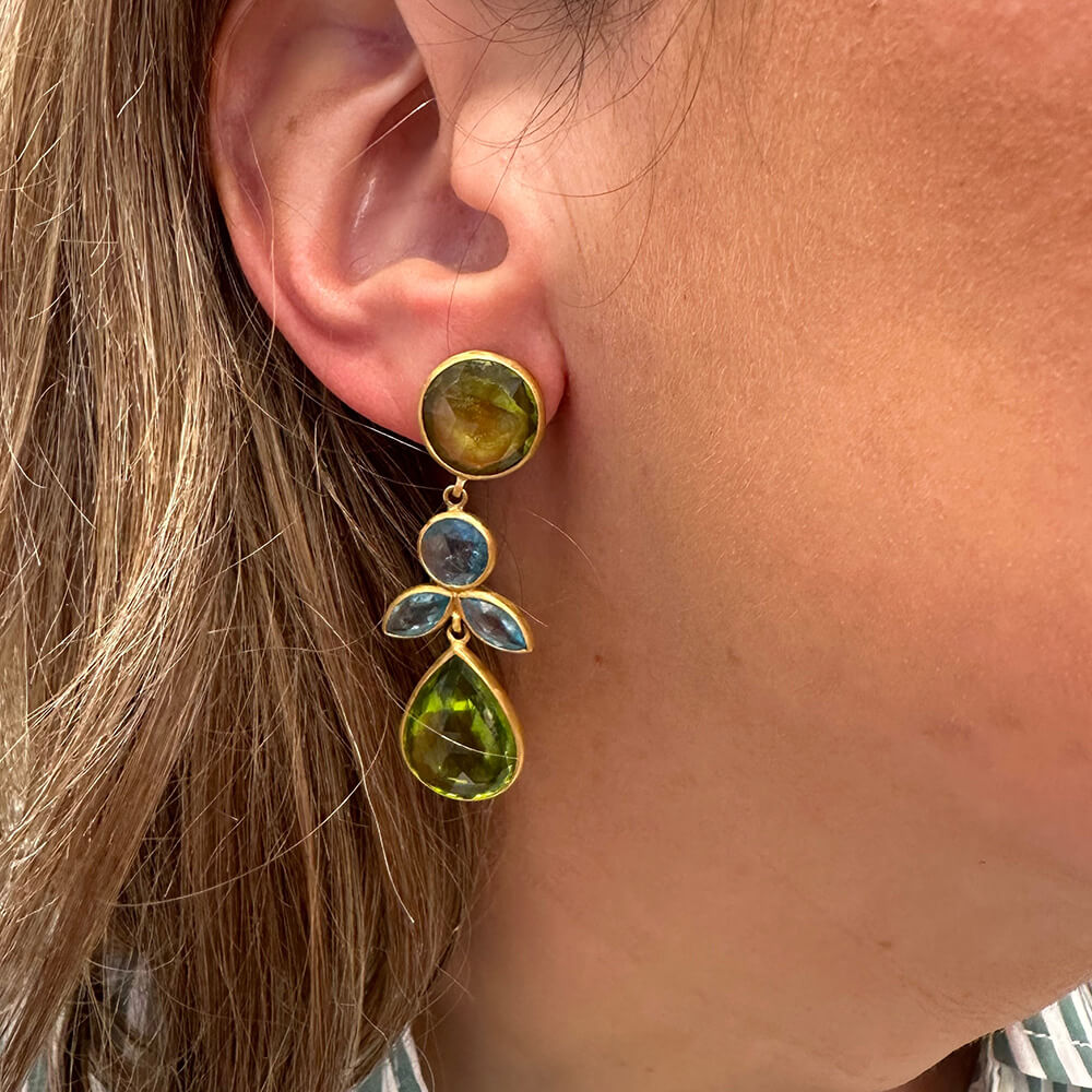 A close-up of a woman's ear wearing a 22k gold  earring with green peridot gemstone accents and small aquamarine stones.