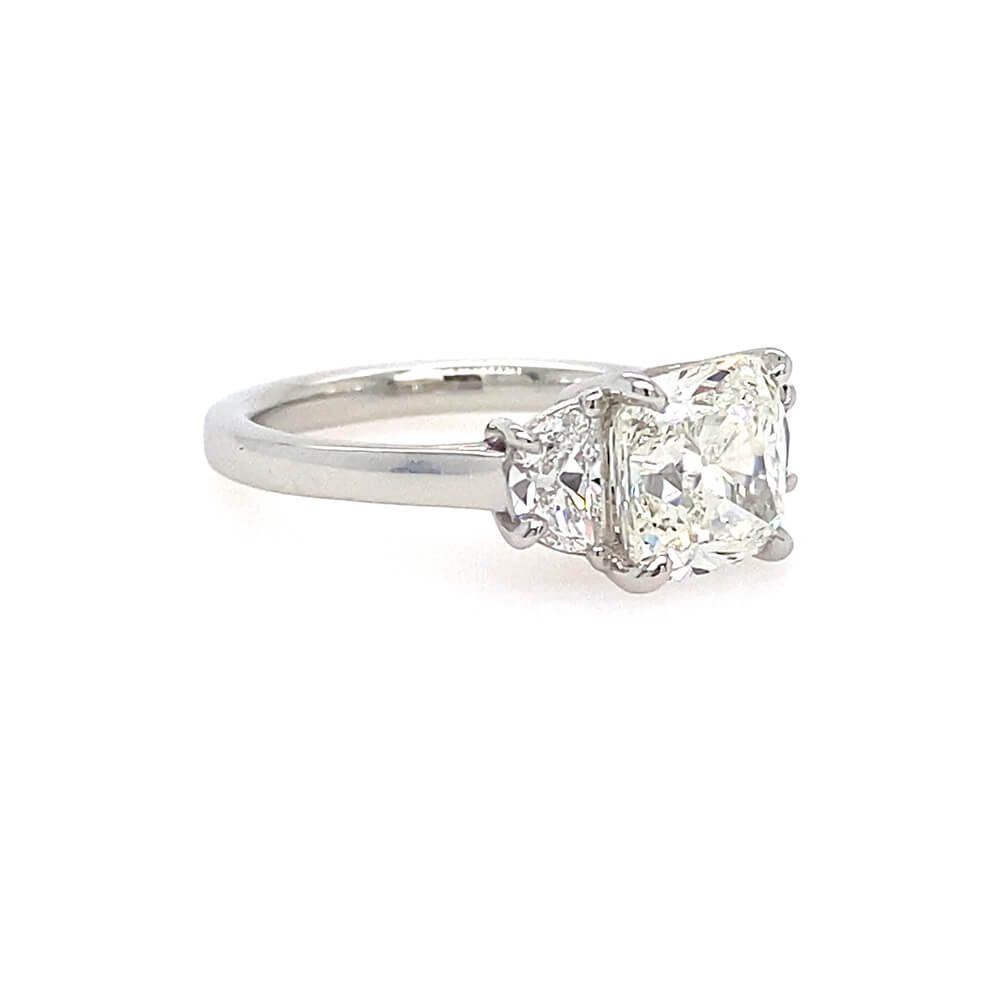 A three-stone diamond engagement ring featuring a cushion-cut center diamond and two half moon side diamonds in a platinum metal band, set against a plain white background.