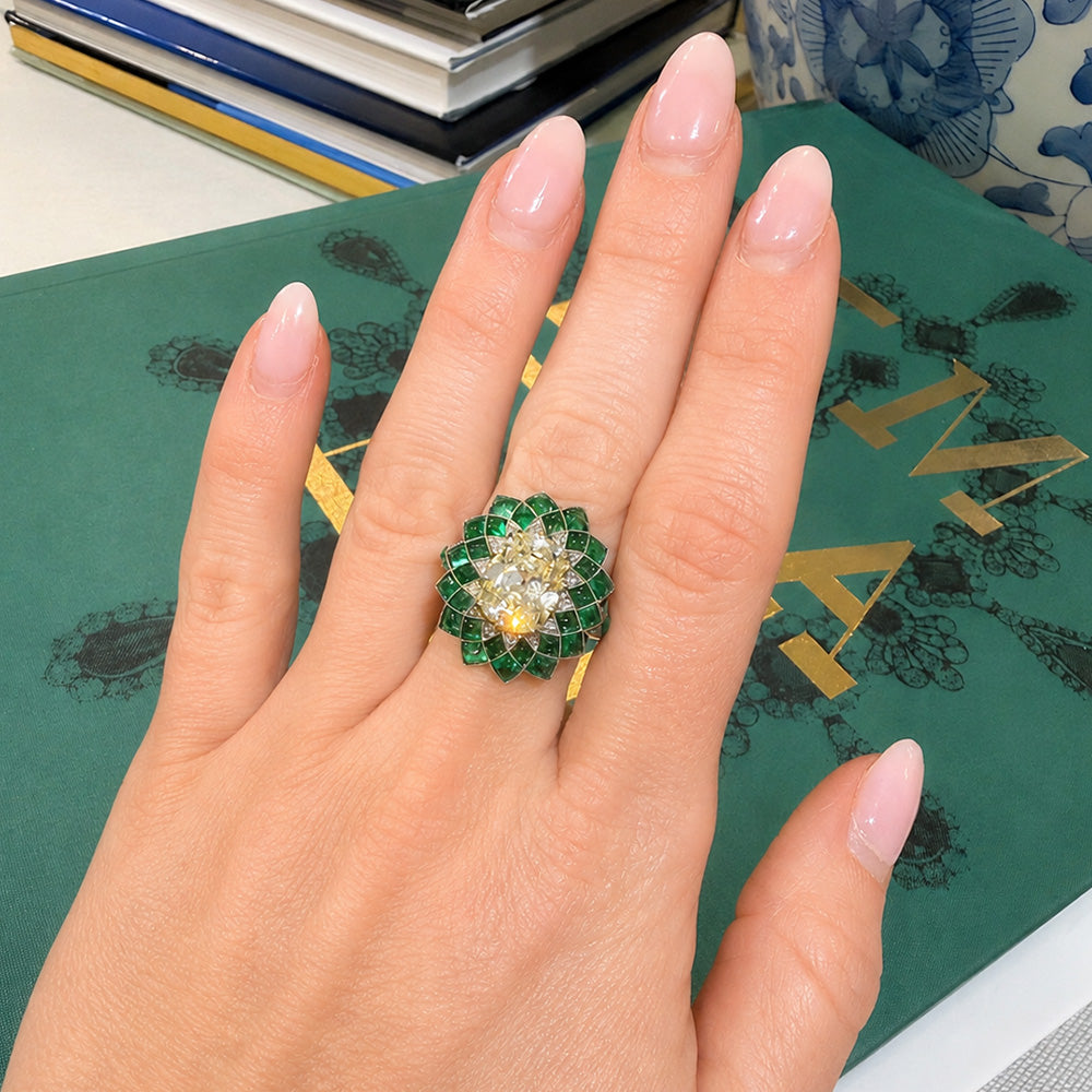 A close-up of a hand wearing a large floral shaped ring with green emerald stones and a central oval diamond 