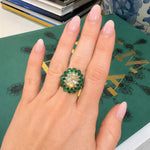 A close-up of a hand wearing a large floral shaped ring with green emerald stones and a central oval diamond 