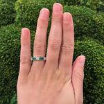 Hand wearing a platinum ring with emerald gemstones and diamonds against a green background