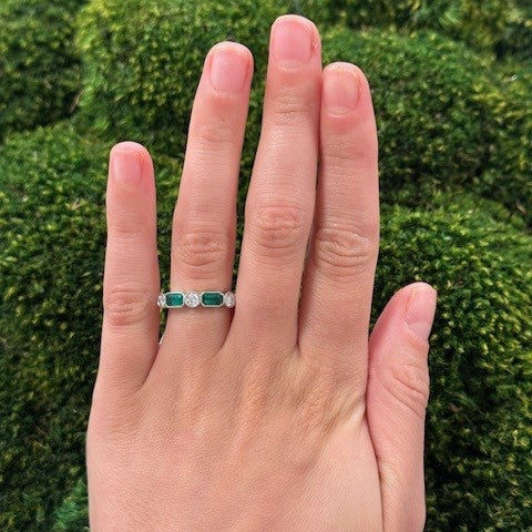 Hand wearing a platinum ring with emerald gemstones and diamonds against a green background