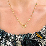 A close-up of a 14K gold chain necklace with a small open circle pendant, worn on a model's neck above a patterned off-the-shoulder top.