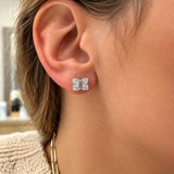 A close-up of a person's ear wearing a square stud earring with a silver-toned setting and clear gemstones.
