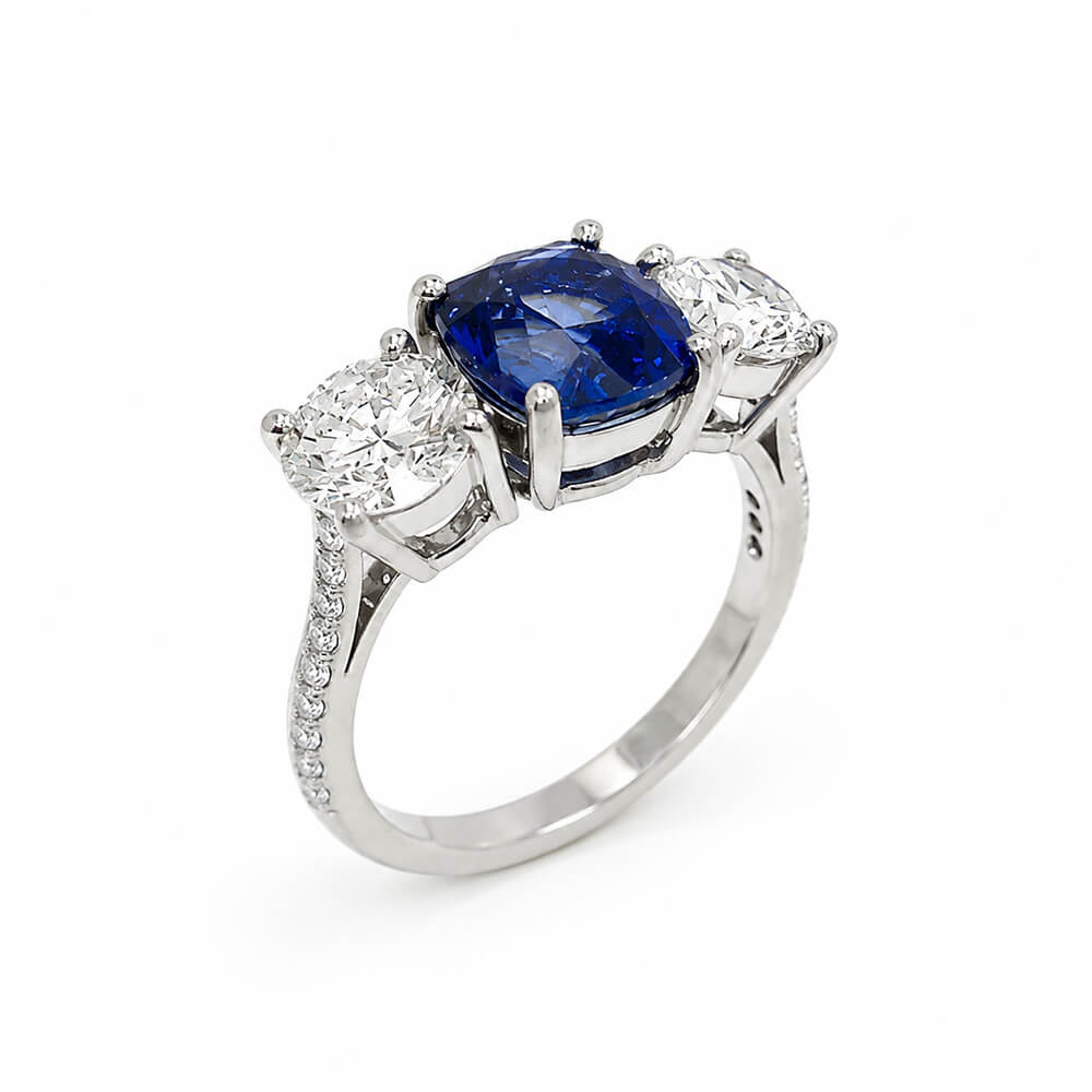A platinum three-stone ring featuring a central cushion-cut deep blue sapphire flanked by two round brilliant-cut diamonds, set on a clean white background.
