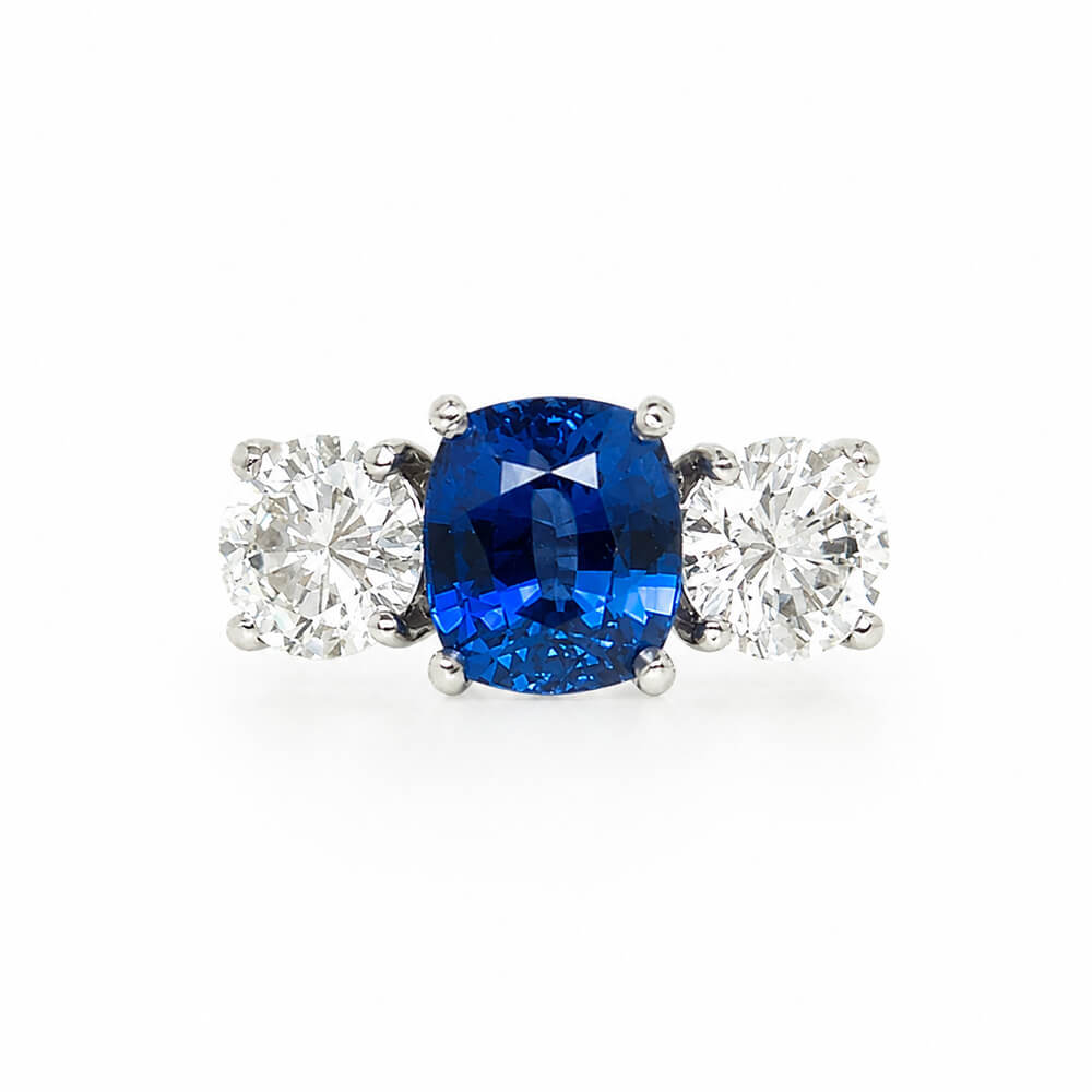 A three-stone ring featuring a central cushion-cut blue sapphire flanked by two round brilliant-cut diamonds in a platinum setting, shown on a plain white background.