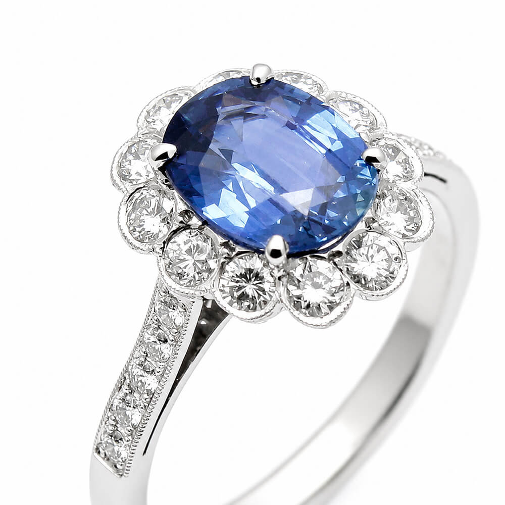 A platinum ring featuring a large oval cushion cut blue sapphire center stone surrounded by a halo of round clear diamonds, with a pavé-set diamond band, shown on a white background.