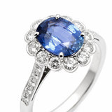 A platinum ring featuring a large oval cushion cut blue sapphire center stone surrounded by a halo of round clear diamonds, with a pavé-set diamond band, shown on a white background.