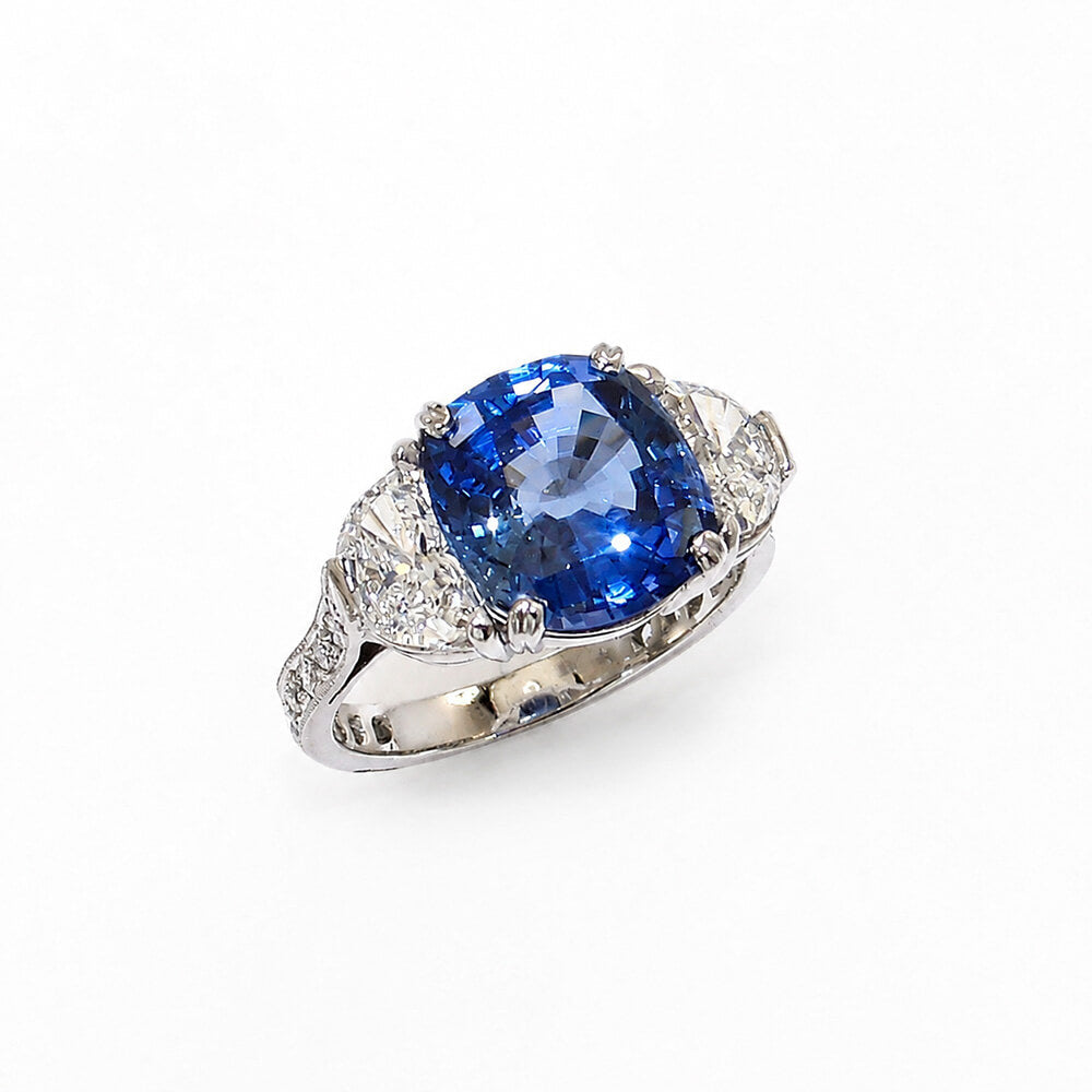 A platinum ring featuring a cushion-cut blue sapphire center stone and smaller clear half moon diamonds on the shoulders, set against a plain white background.
