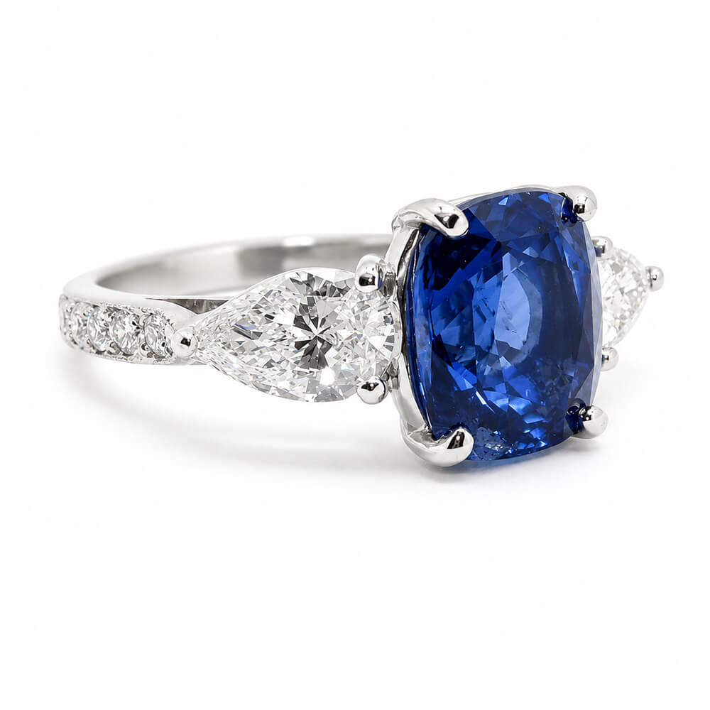 A platinum ring featuring a large cushion-cut blue sapphire center stone, flanked by two pear shaped clear diamonds and a smaller round diamond, set on a white background.