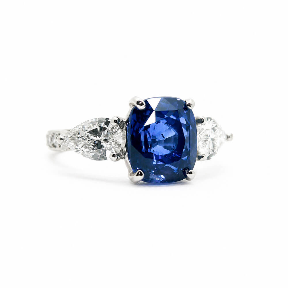 A ring featuring a cushion-cut blue sapphire center stone flanked by two pear shapedclear diamonds on a platinum band, set against a plain white background.