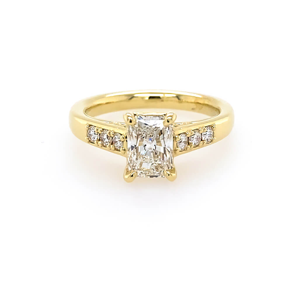An 18K yellow gold engagement ring featuring a rectangular radiant-cut diamond center stone and smaller round diamonds set along the shoulders, shown on a plain white background.