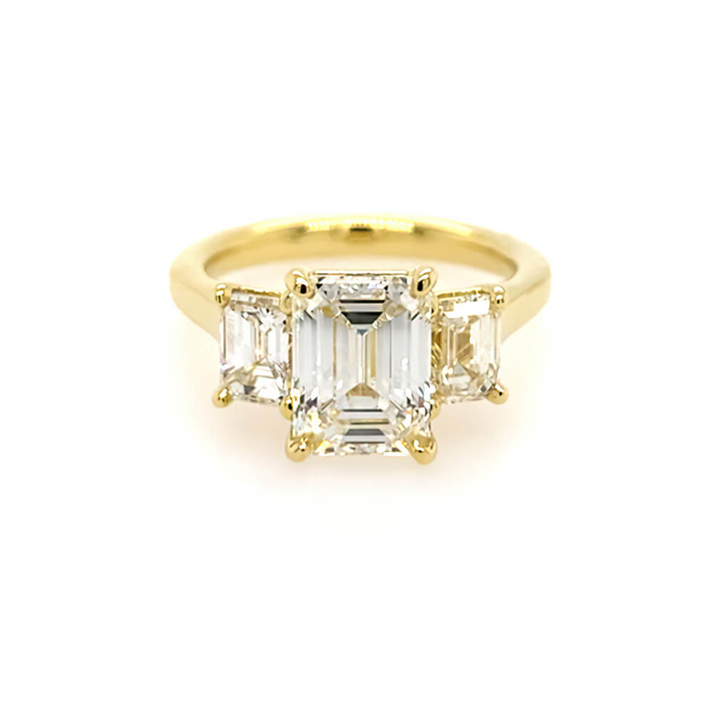 An 18K yellow gold three-stone ring featuring a large emerald-cut center diamond flanked by two smaller emerald-cut side diamonds, set against a plain white background.