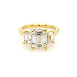An 18K yellow gold three-stone ring featuring a large emerald-cut center diamond flanked by two smaller emerald-cut side diamonds, set against a plain white background.