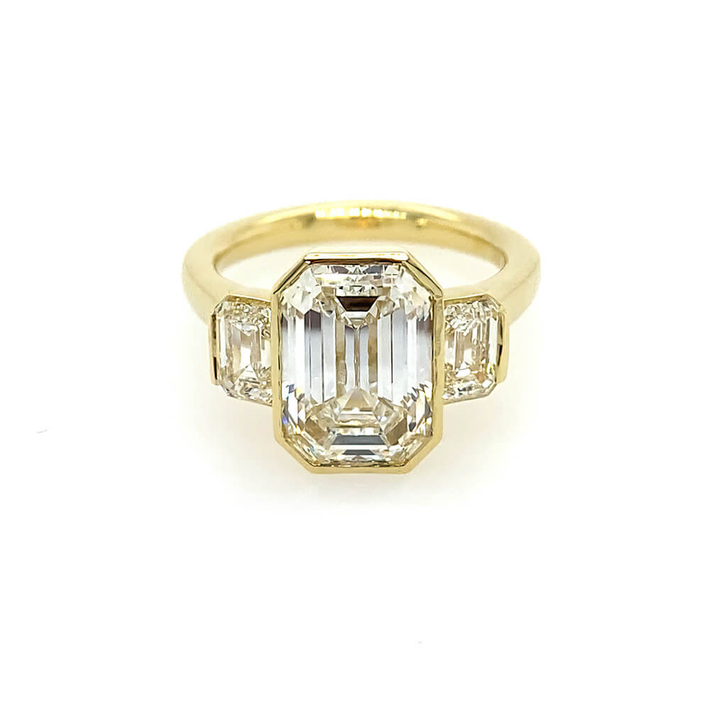An 18K yellow gold ring featuring a large emerald-cut center stone with two smaller emerald-cut side stones, set against a plain white background.