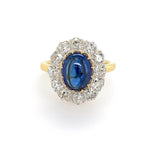 A vintage-style ring featuring an oval-cut blue sapphire surrounded by a halo of clear diamonds on an 18K yellow gold band, set against a plain white background.