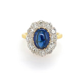 A vintage-style ring featuring an oval-cut blue sapphire surrounded by a halo of clear diamonds on an 18K yellow gold band, set against a plain white background.