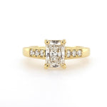An 18K yellow gold engagement ring featuring a large rectangular radiant-cut center diamond flanked by smaller round diamonds on the band, set against a plain white background.