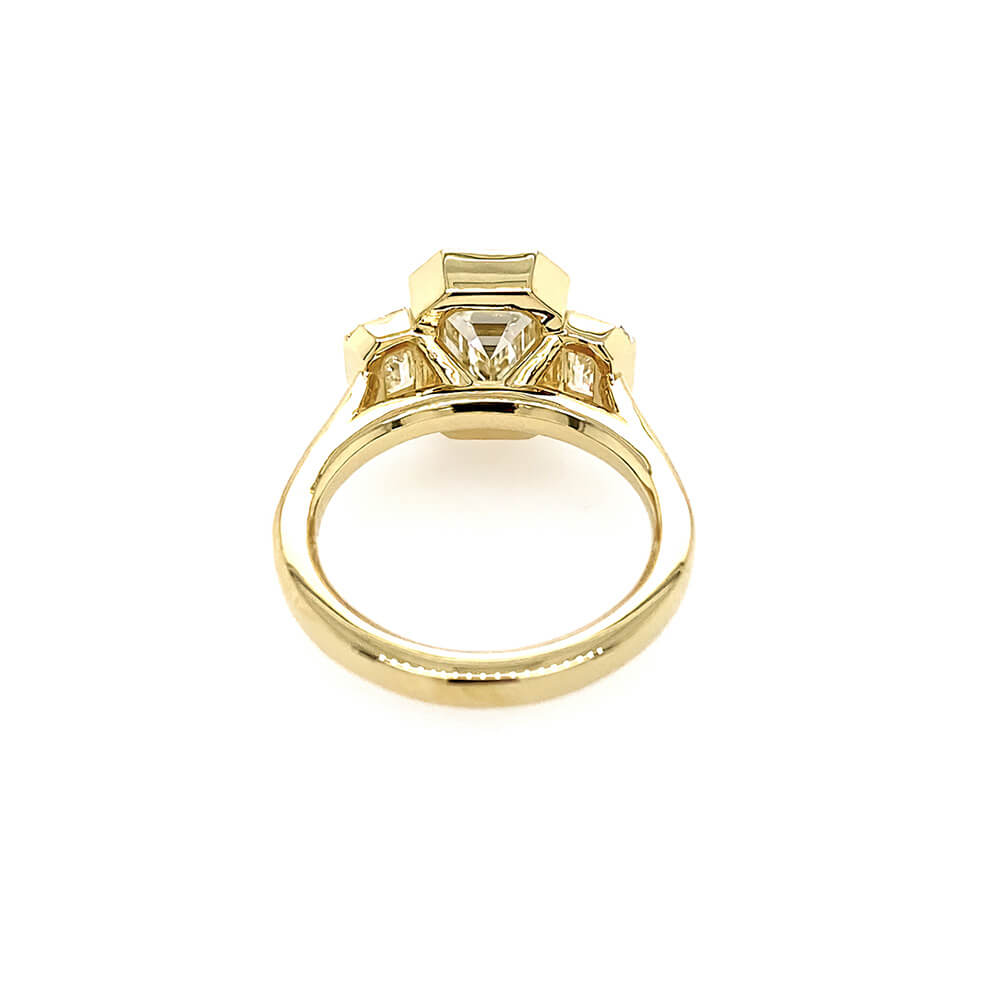 An 18K yellow gold ring featuring a central emerald-cut diamond and smaller side diamonds, shown from a top-down view on a plain white background.