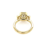An 18K yellow gold ring featuring a central emerald-cut diamond and smaller side diamonds, shown from a top-down view on a plain white background.