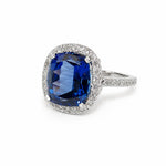 A cushion-cut blue sapphire ring with a diamond halo and a platinum pavé diamond band, set against a plain white background.