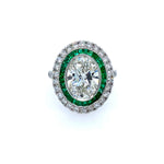 A ring with a diamond center stone surrounded by emeralds and a diamond border, set in a platinum band.