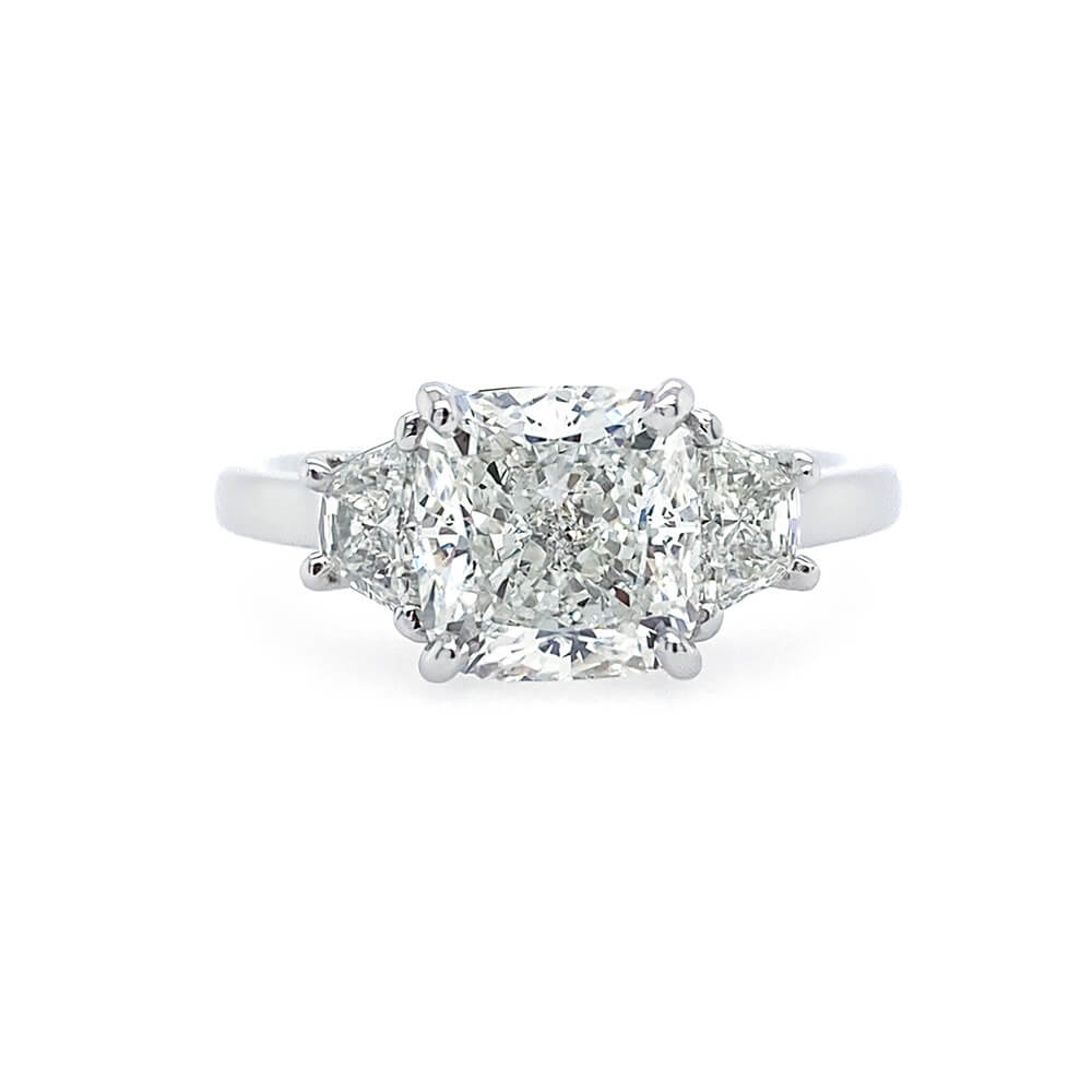 A three-stone diamond ring featuring a central cushion-cut diamond flanked by two smaller half moon diamonds on a polished platinum band, set against a plain white background.