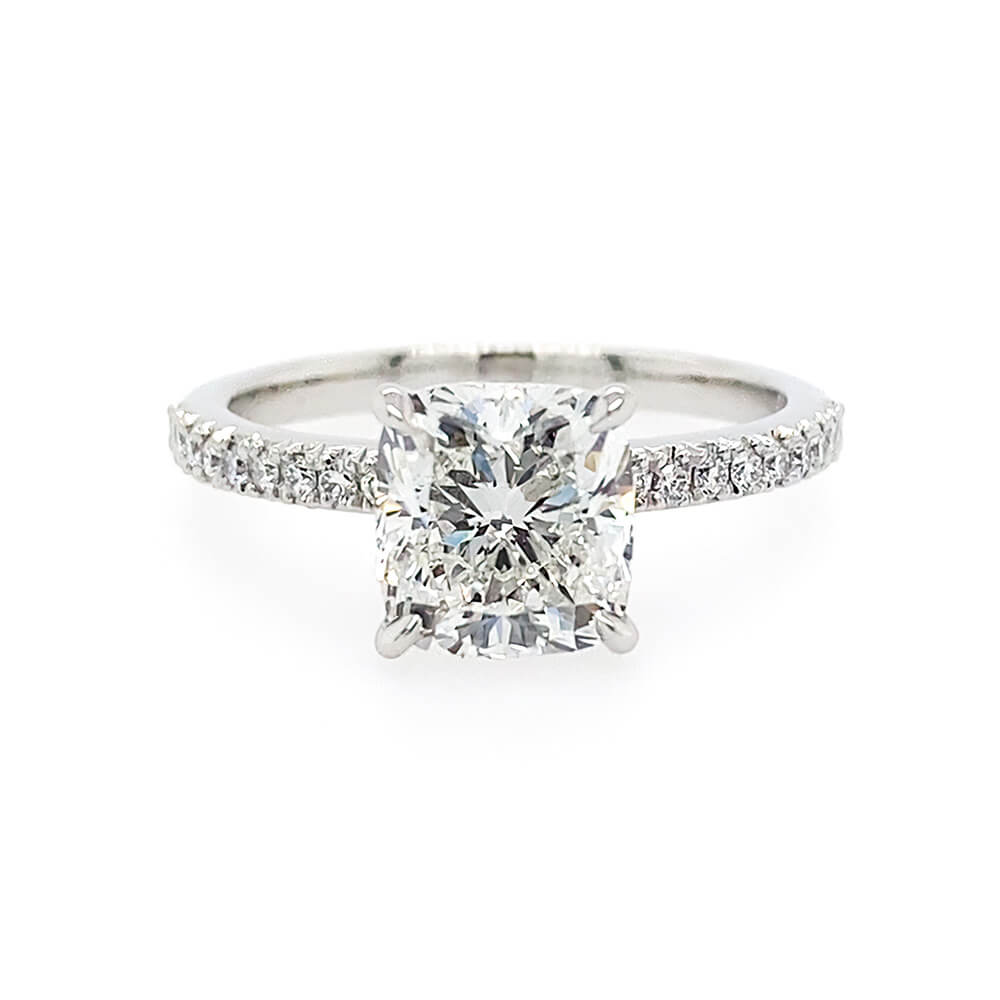 A square cushion cut diamond engagement ring with a platinum band set with small pavé diamonds, shown on a plain white background.
