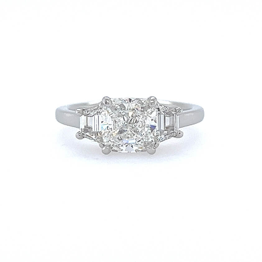 A three-stone diamond ring featuring a central cushion-cut diamond flanked by two half-moon-cut diamonds on a polished platinum band, set against a plain white background.