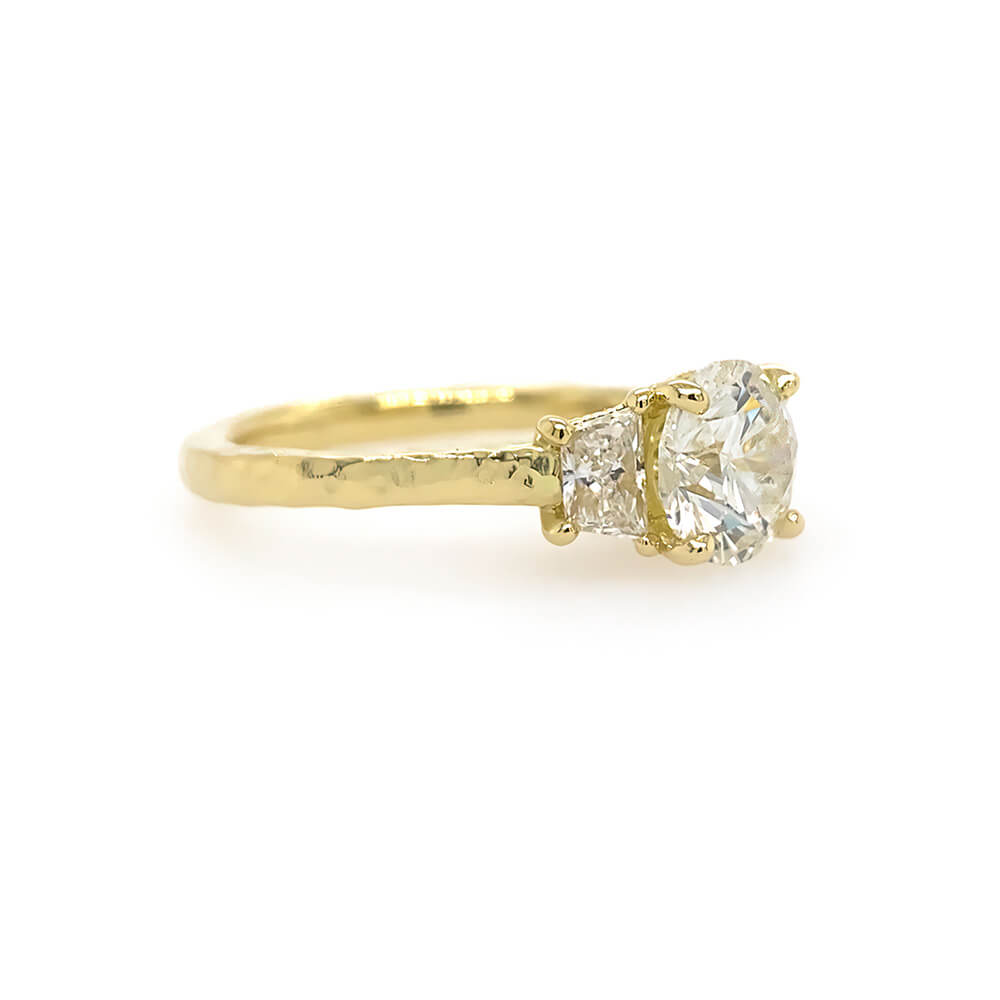 A custom designed round brilliant diamond ring with trapezoid side diamonds in hammered 18K yellow gold