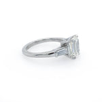 A platinumsolitaire engagement ring featuring an emerald cut center stone in a four-prong setting, with two baguette side stones, shown on a plain white background.