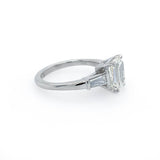 A platinumsolitaire engagement ring featuring an emerald cut center stone in a four-prong setting, with two baguette side stones, shown on a plain white background.
