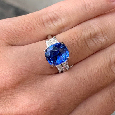 A close-up of a hand wearing a platinum ring with a large cushion-cut blue sapphire and small clear accent diamonds on the band, set against a blurred outdoor background.