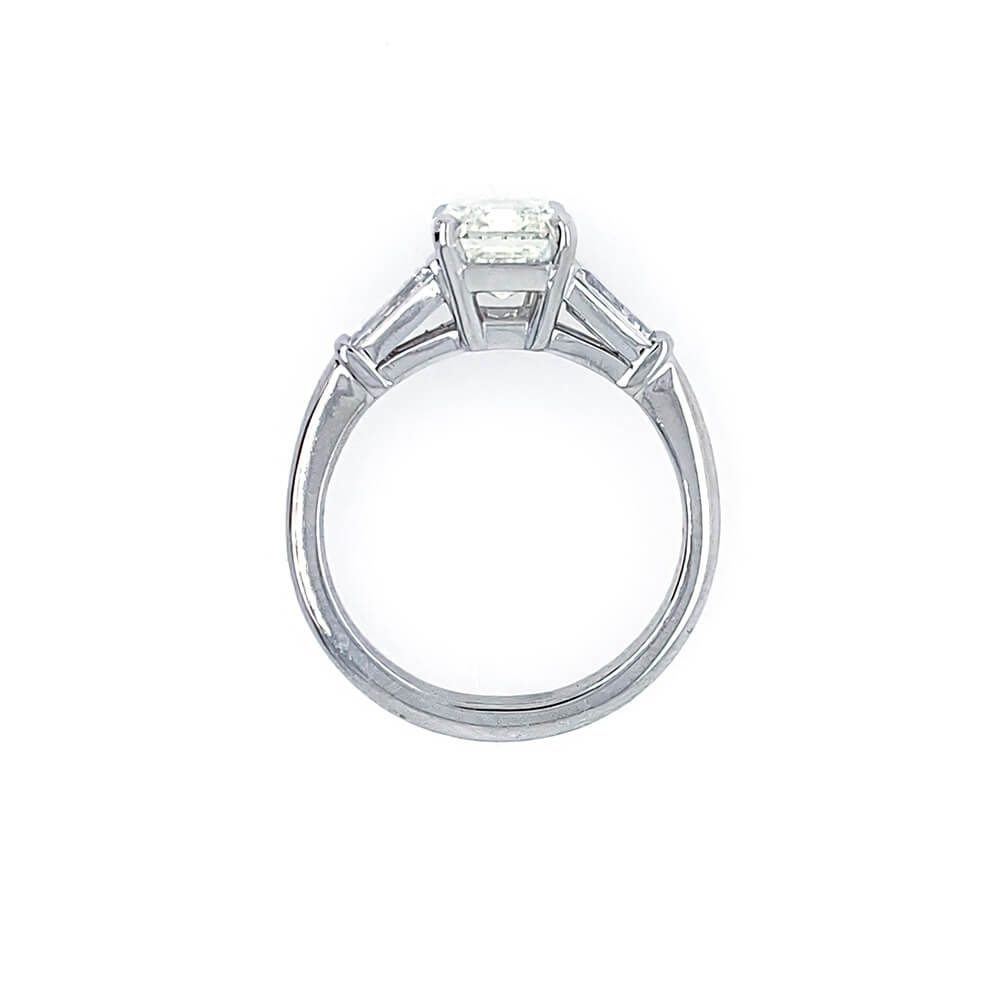A platinumsolitaire engagement ring featuring an emerald cut center stone in a four-prong setting, with two baguette side stones, shown on a plain white background.