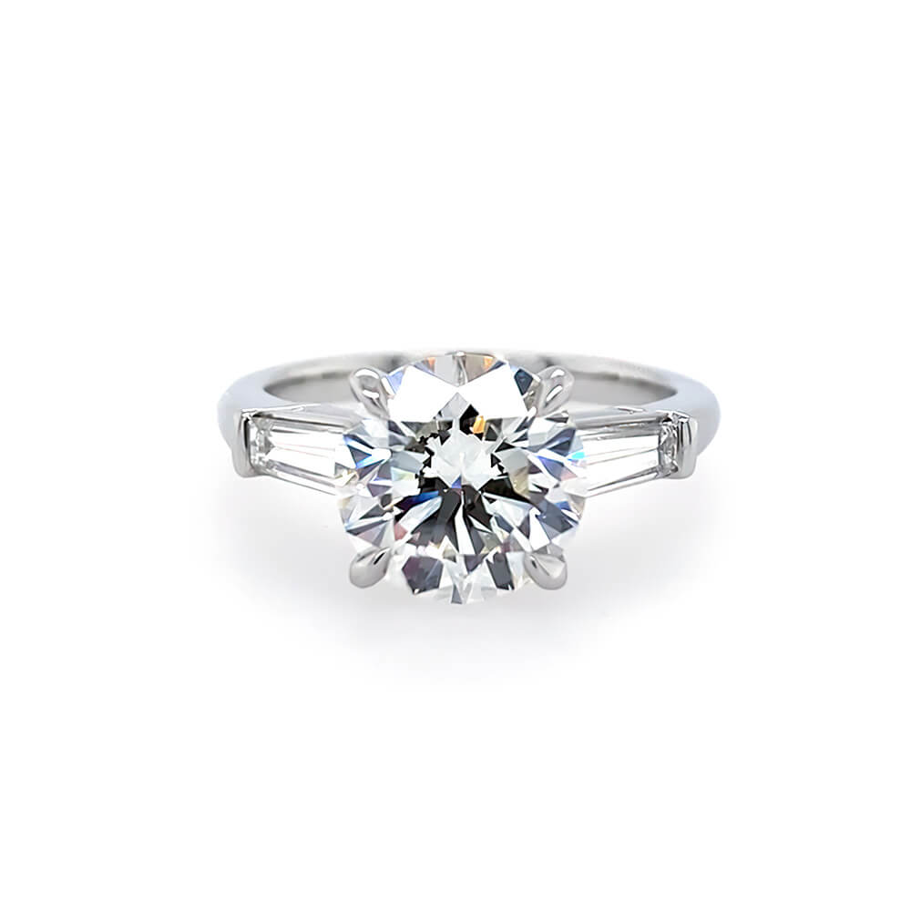 A classic solitaire engagement ring featuring a round brilliant-cut center diamond in a four-prong setting, flanked by two baguette-cut side stones on a polished platinum band, set against a plain white background.