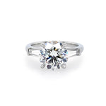 A classic solitaire engagement ring featuring a round brilliant-cut center diamond in a four-prong setting, flanked by two baguette-cut side stones on a polished platinum band, set against a plain white background.