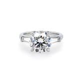 A classic solitaire engagement ring featuring a round brilliant-cut center diamond in a four-prong setting, flanked by two baguette-cut side stones on a polished platinum band, set against a plain white background.