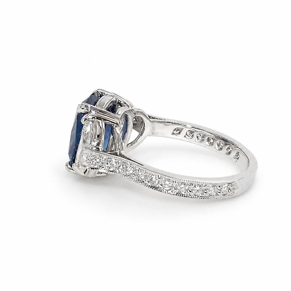 A platinum ring featuring a cushion-cut blue sapphire center stone and smaller clear half moon diamonds on the shoulders, set against a plain white background.