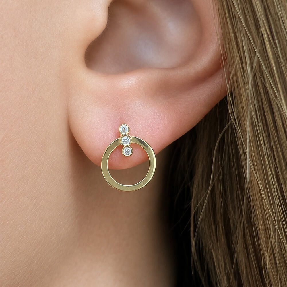 A close-up of a woman's ear wearing a gold hoop earring with three small clear diamonds stacked vertically on the post.